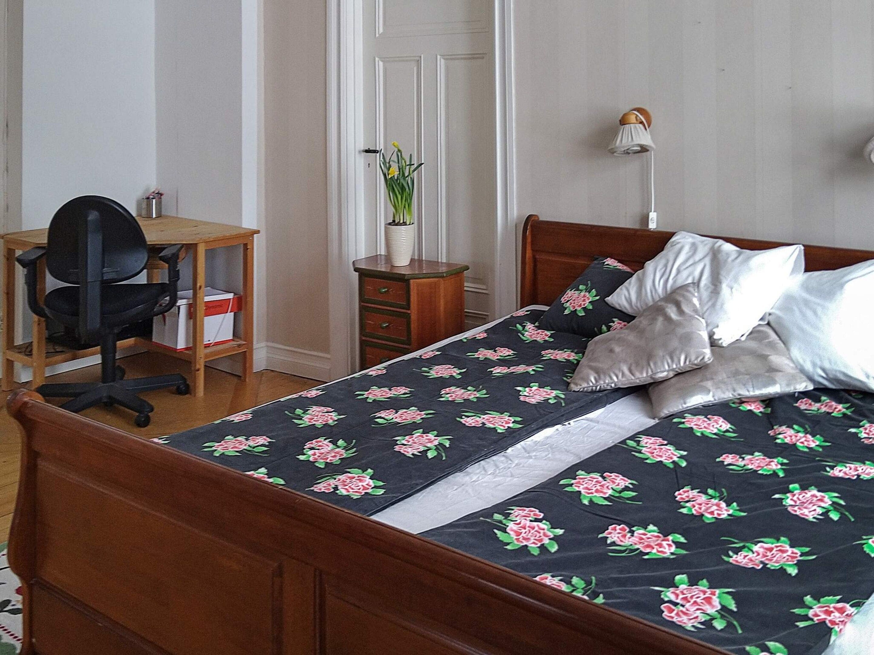 A bedroom with a wooden bed, a desk with a chair, a bedside table, and a lamp.