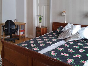 A bedroom with a wooden bed, a desk with a chair, a bedside table, and a lamp.