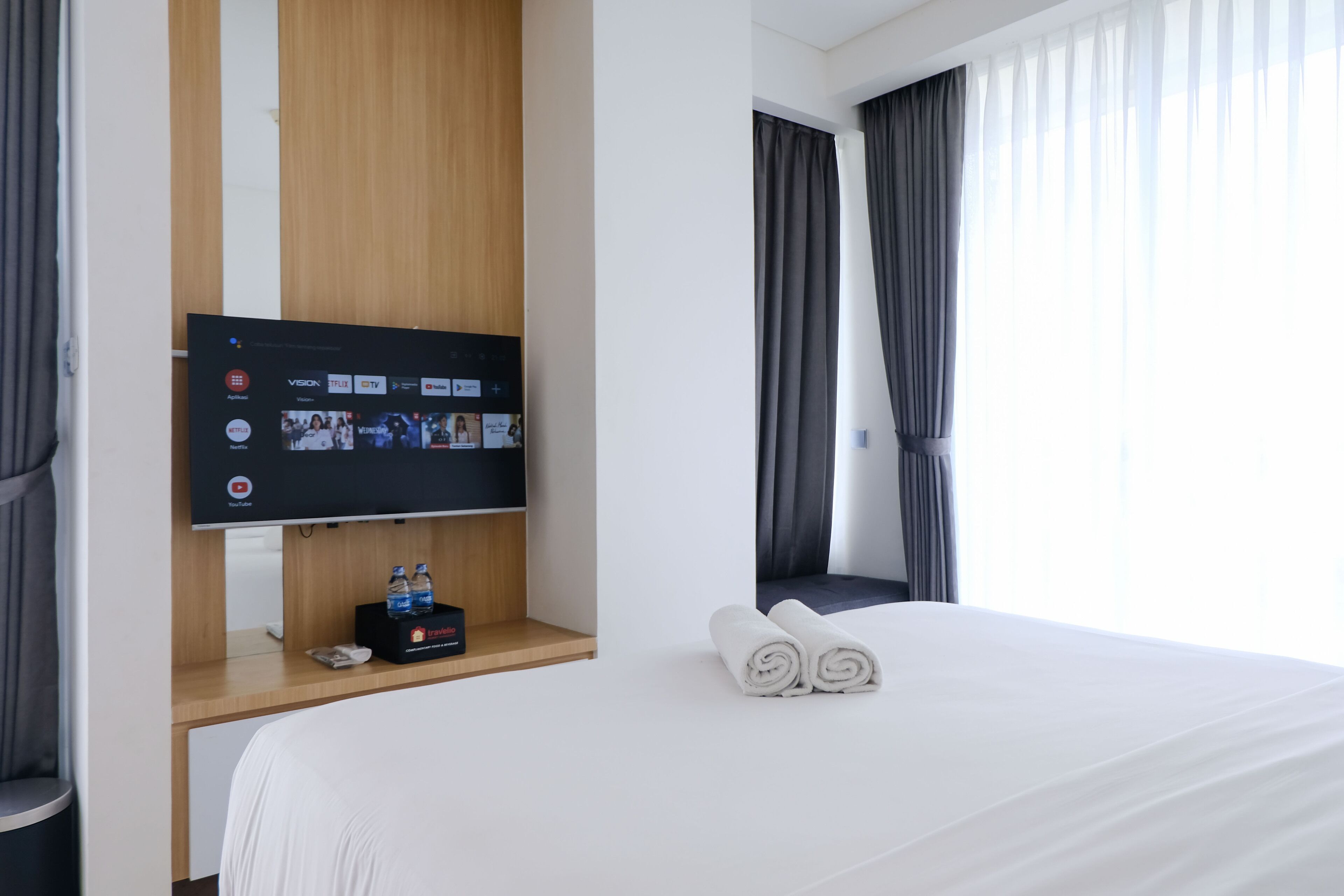 Una habitación de hotel con una cama, un televisor de pantalla plana montado en un estante de madera, una botella de agua y una toalla enrollada sobre la cama.