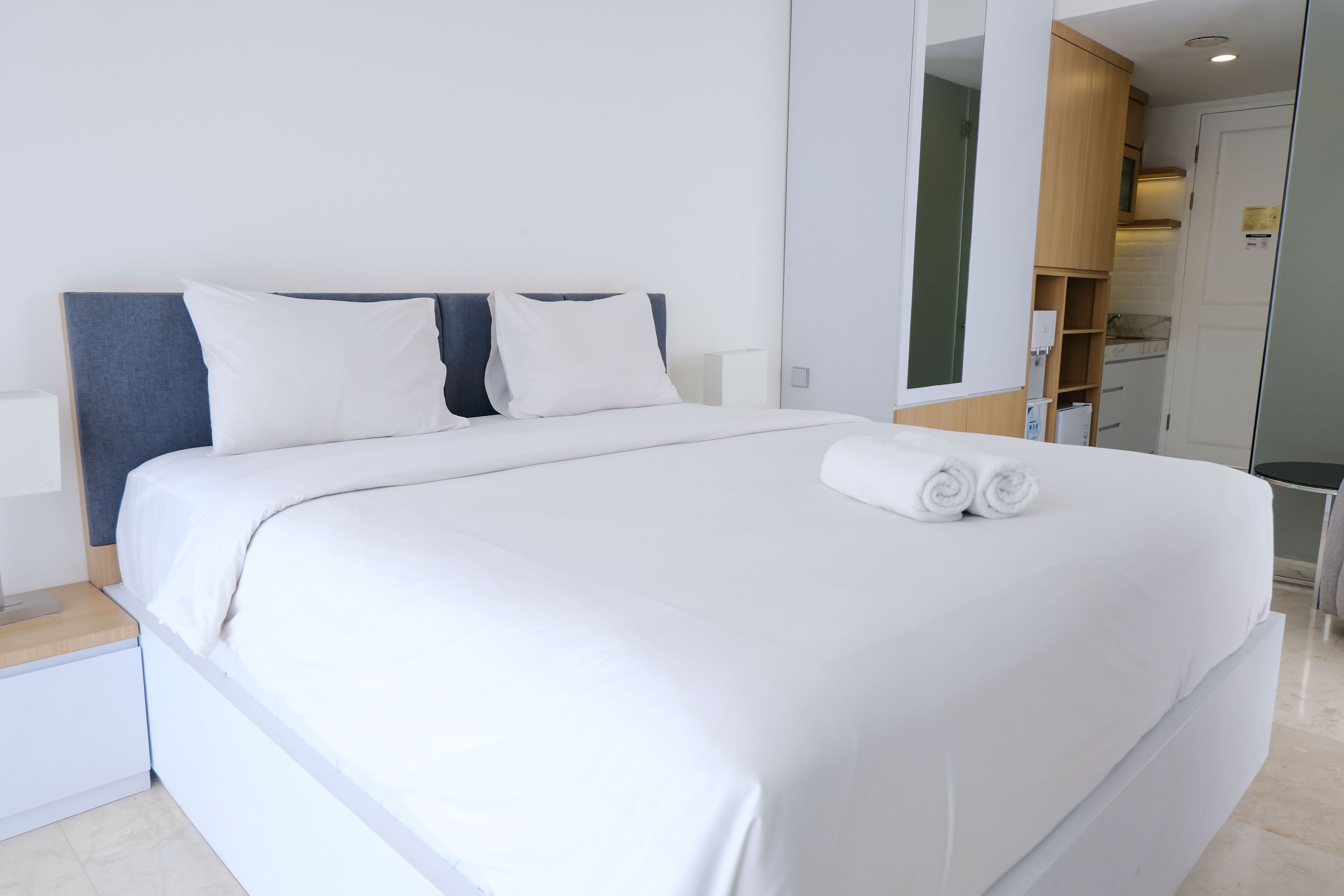 Una cama bien hecha con sábanas blancas y dos almohadas, una mesita de noche de madera y una pared blanca.