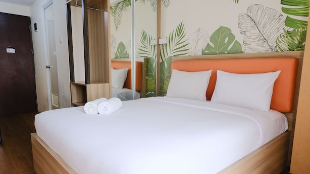 Un lit bien fait, avec du linge de lit blanc et une tête de lit orange, un plancher en bois et un mur orné d’un motif de feuilles tropicales.