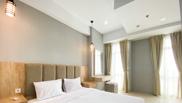 A modern hotel room with a large bed, wooden headboard, bedside tables, a nightstand, a mirror, and a window with curtains. - Simply Look And Comfort 1Br Vasanta Innopark Apartment (Cikarang)