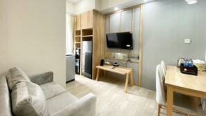 A compact living space with a sofa, a dining table with chairs, a TV mounted on a wooden panel, a refrigerator, and a coffee maker. - Simply Look And Comfort 1Br Vasanta Innopark Apartment (Cikarang)
