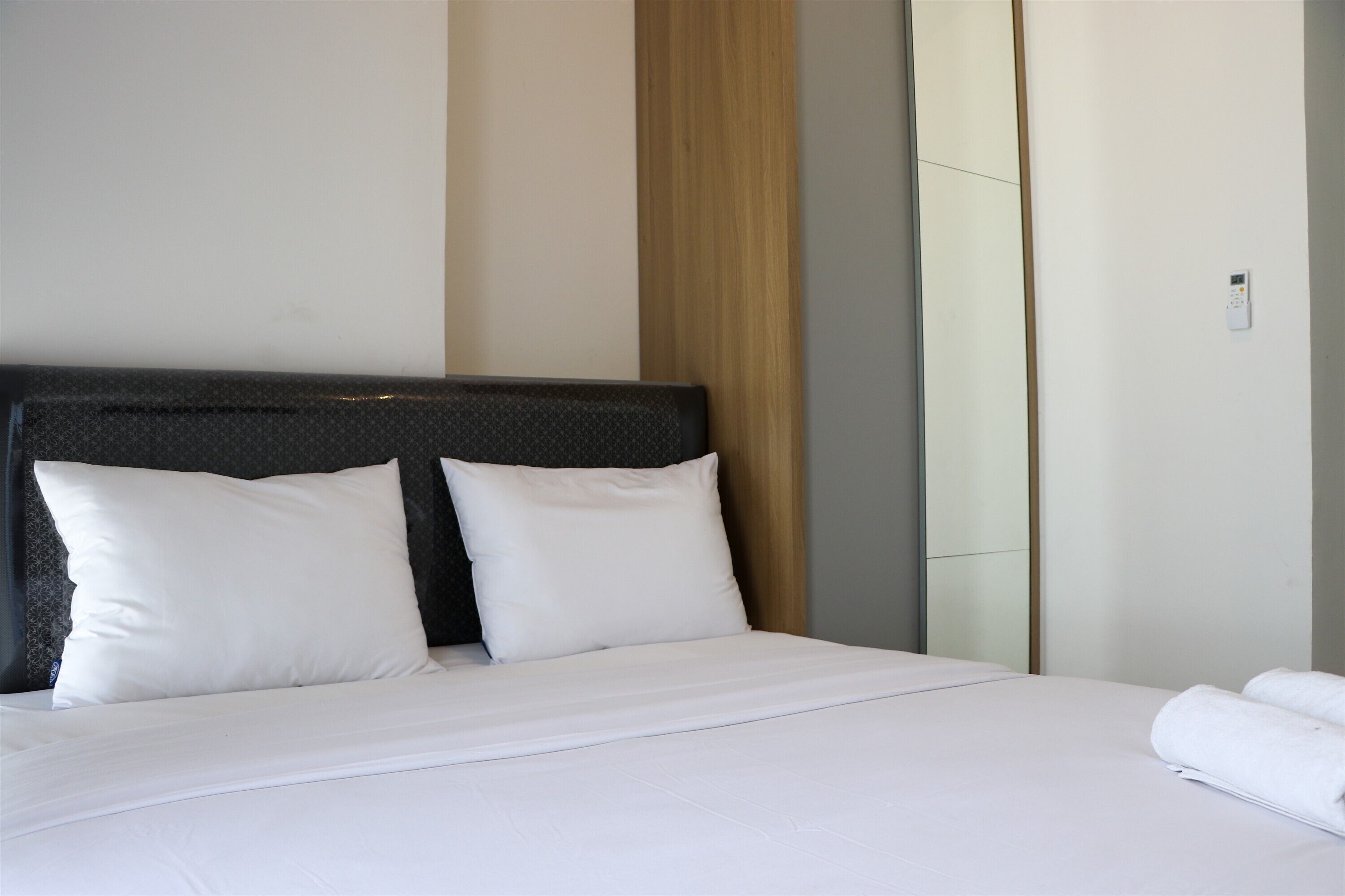 A neatly made bed with white linens and pillows, a wooden headboard, and a wall-mounted thermostat.