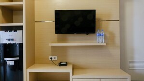 Ein Hotelzimmer mit Minibar, einem flachbildfernseher an der Wand, einem Wasserspender und einem kleinen Regal mit Wasserflaschen.