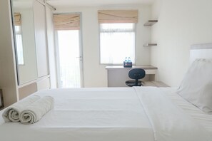 Et hotelværelse med en pænt redt seng, et skrivebord med stol, et spejl og et vindue med gardiner.