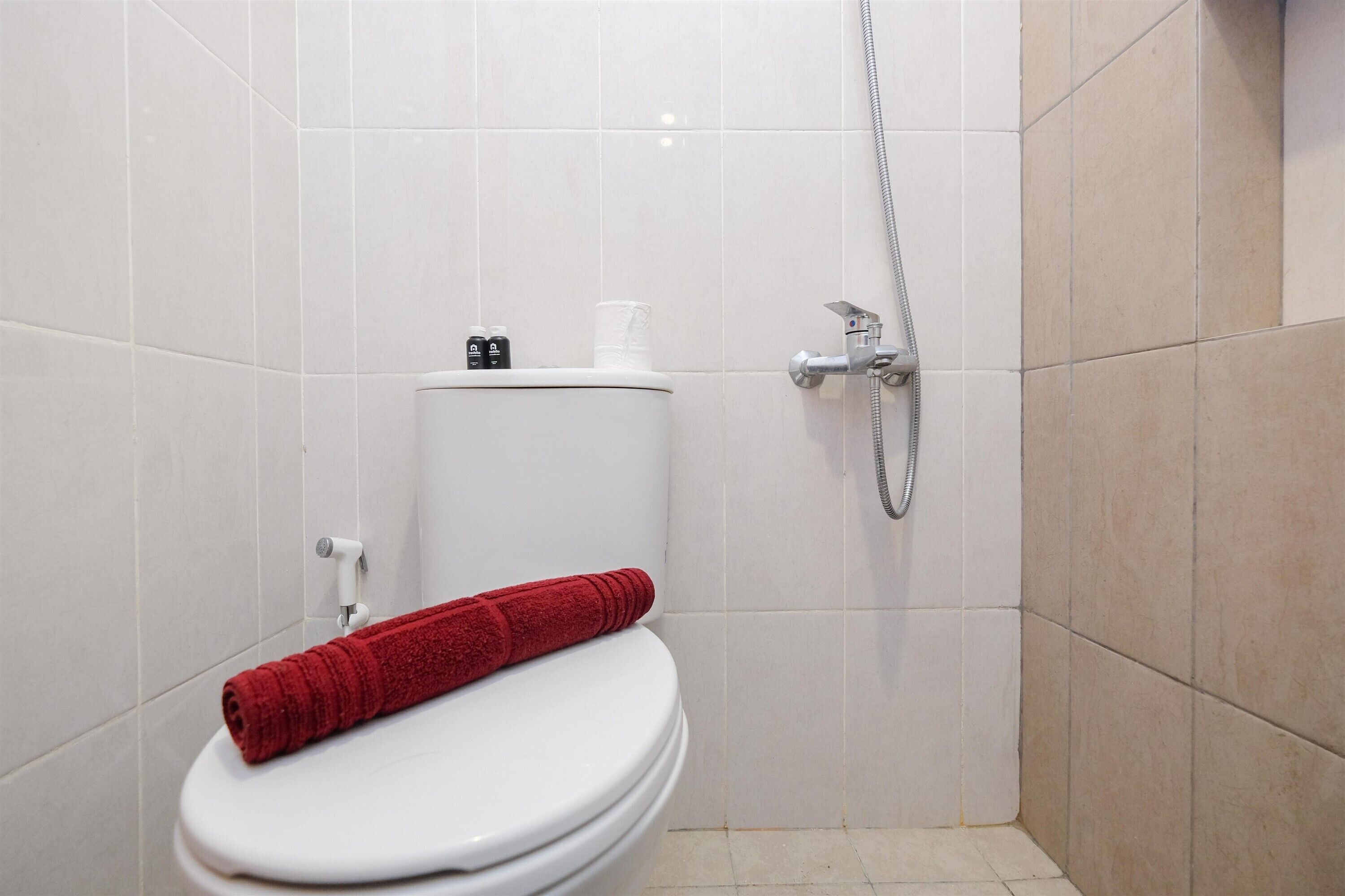 A bathroom with a white toilet, a red towel on the seat, a shower head, and a toilet paper holder.