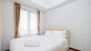A neatly made bed with white linens and a rolled white towel, a wooden headboard, and beige curtains on a window.
