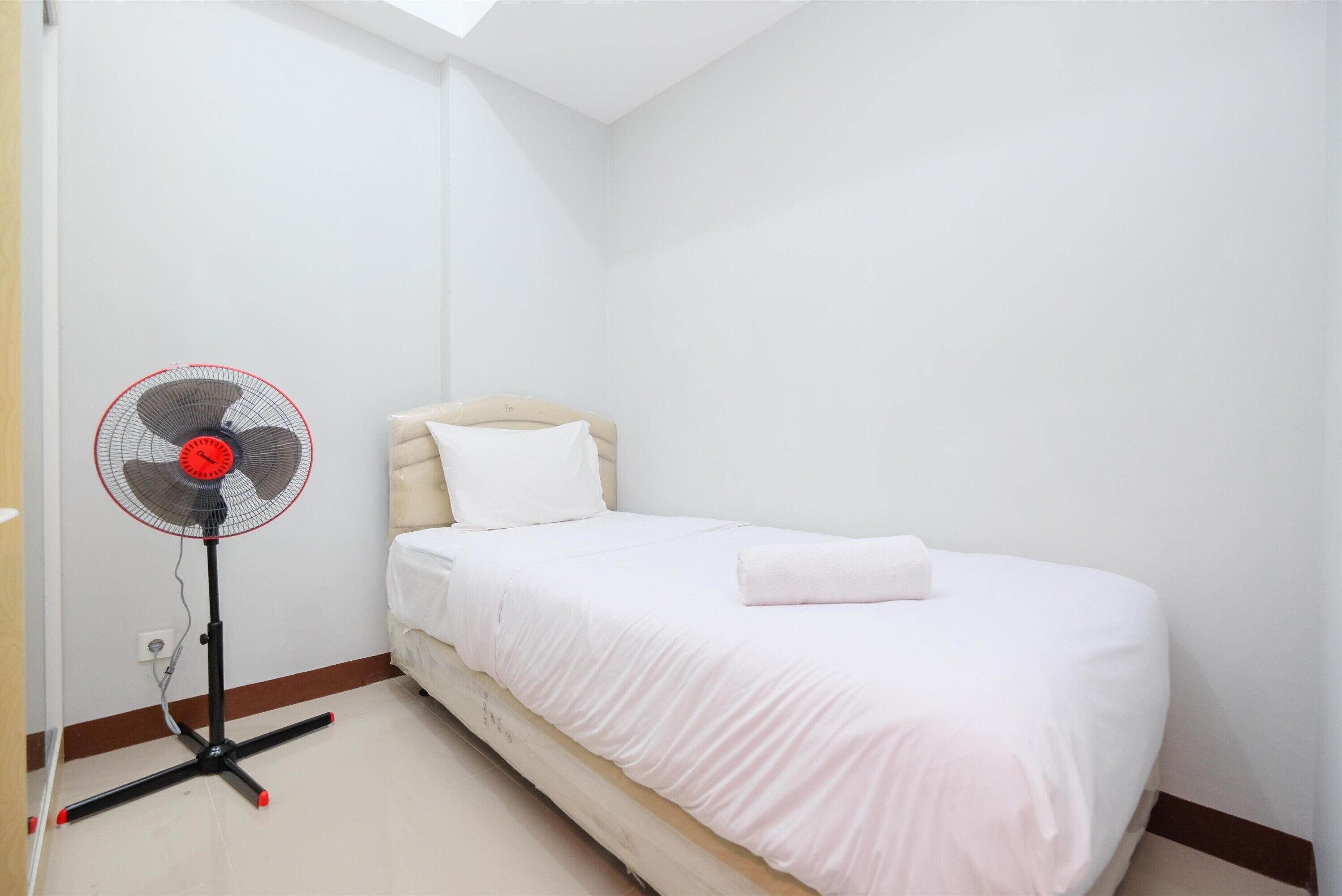 A small, neatly made room with a single bed, a standing fan, and a white wall.