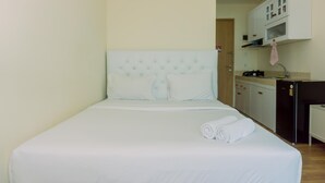 A neatly made bed with white linens and pillows, a small kitchen area with a refrigerator and cabinets, and a door leading to another room. - Good Deal And Simply Look Studio Apartment At B Residence (Pagedangan)
