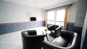 Ein Zimmer mit Fliesenboden, zwei Ledersessel, einem kleinen Glastisch, einem an der Wand montierten Fernseher und einem großen Fenster mit Vorhängen.
