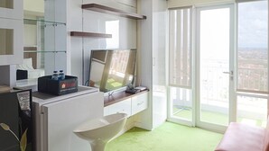 A modern hotel room with a large TV, a minibar, a glass shower enclosure, and a balcony with a city view. - Comfort 1Br At Vida View Makassar Apartment (Makassar)