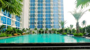 A swimming pool with a view of a high-rise building, surrounded by palm trees and greenery. - Strategic And Well Designed Studio Apartment At Capitol Park Residence (Jakarta)