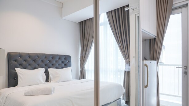 A modern hotel room with a tufted headboard, a large bed with white linens, a glass door leading to a balcony, and a large window with curtains. - Strategic And Well Designed Studio Apartment At Capitol Park Residence (Jakarta)