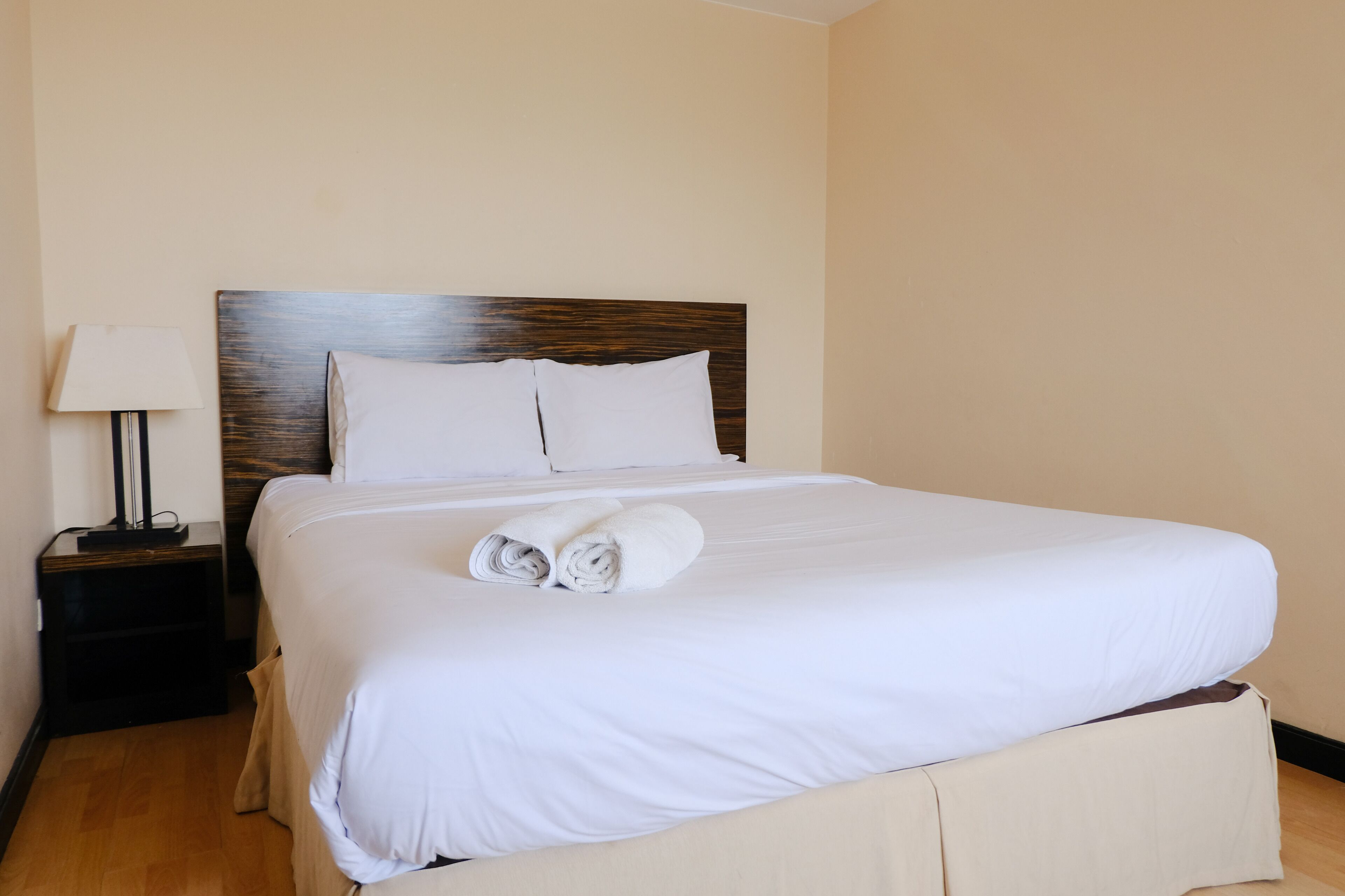 A neatly made bed with white linens and two rolled towels, a bedside lamp, and a wooden headboard in a hotel room.
