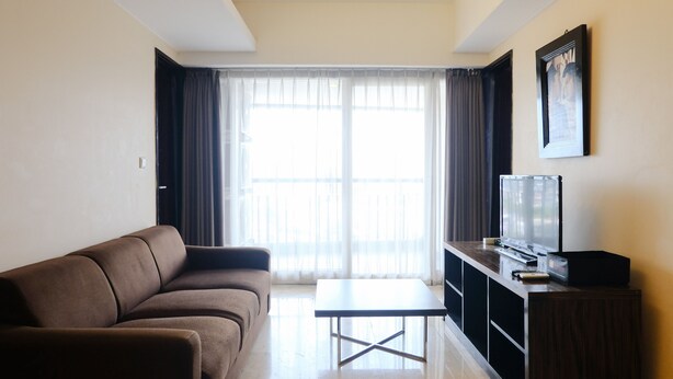 A living room with a brown sofa, a coffee table, a TV on a wooden stand, and a large window with sheer curtains. - Homey 2Br At Braga City Walk Apartment (Bandung)