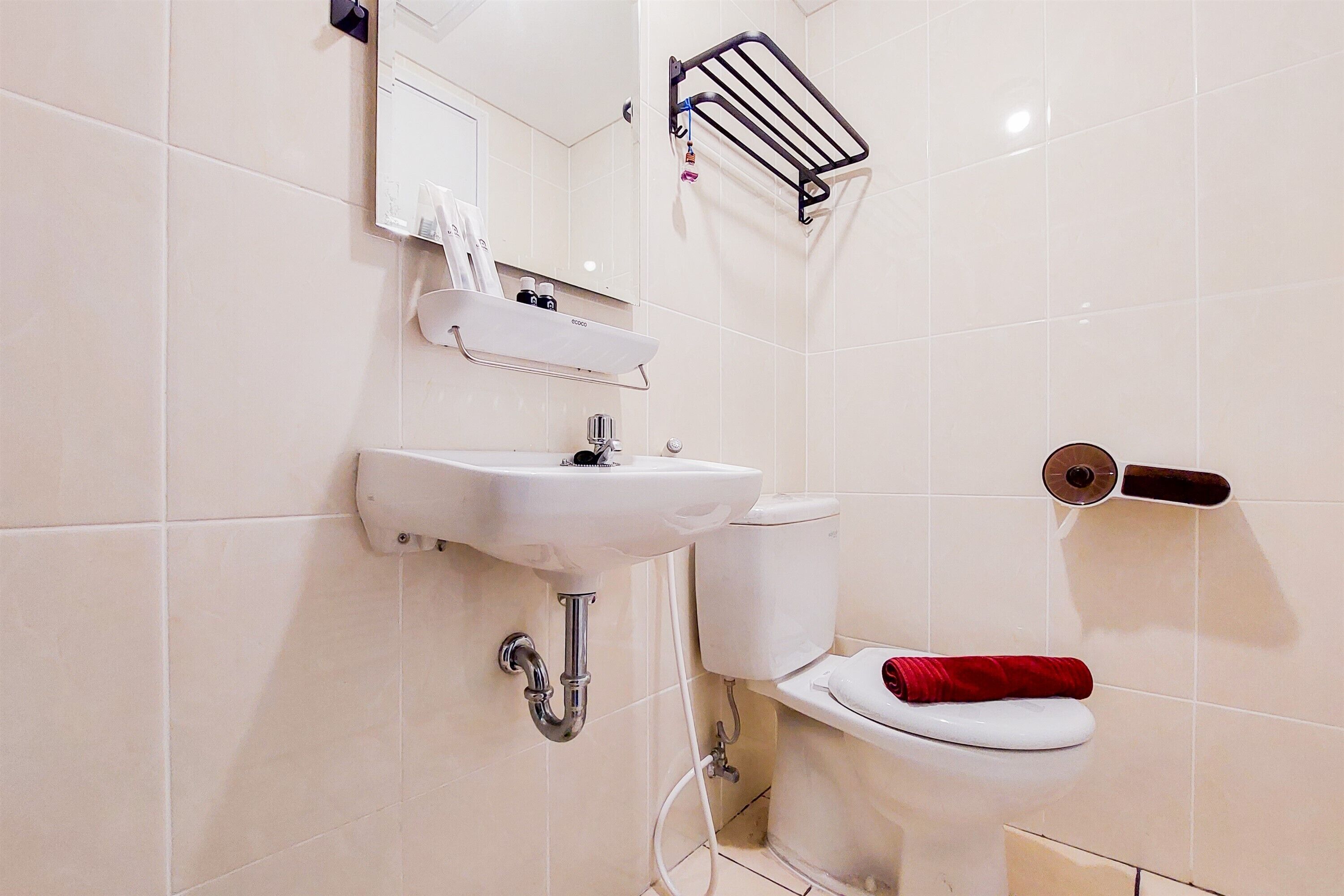 Une salle de bain avec un lavabo, des toilettes et un miroir, tous de couleur blanche. On trouve un porte-serviettes au-dessus du lavabo et un support à papier toilette sur le mur.