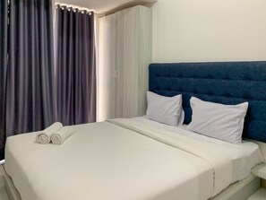 Une chambre d’hôtel avec un lit, du linge de lit blanc, une tête de lit bleue et des rideaux.