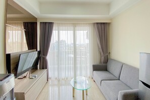 A modern living room with a flat-screen TV, a glass coffee table, a grey sofa, and sheer curtains.