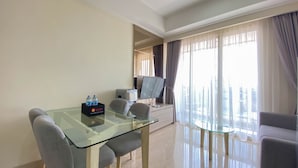 A modern living room with a glass dining table, grey upholstered chairs, a flat-screen TV, and a city view through large windows. - Nice And Minimalist 2Br At Menteng Park Apartment (Jakarta)