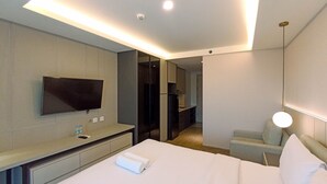 Habitación de hotel con cama, televisor montado en la pared, escritorio con silla y una mesita de noche con lámpara.