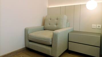 A small, modern room with a light-colored armchair, a bedside table with drawers, and a wall-mounted light fixture.