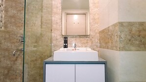 A modern bathroom with a glass shower enclosure, a white sink with a mirror above, and a cabinet below. - Cozy Studio Room At Mataram City Apartment (Ngaglik)