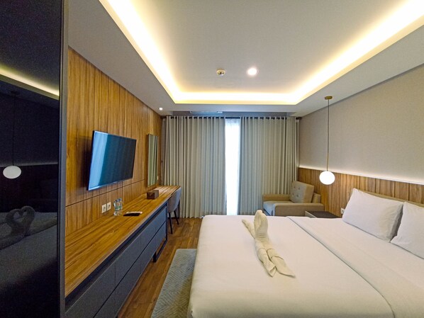 A modern hotel room with a wooden accent wall, a flat-screen TV, a desk with a chair, a bed with a towel, and a large window with curtains.