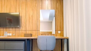 A modern room with a wooden wall, a flat-screen TV mounted above a desk, a grey chair, and a white lamp. - Cozy Studio Room At Mataram City Apartment (Ngaglik)
