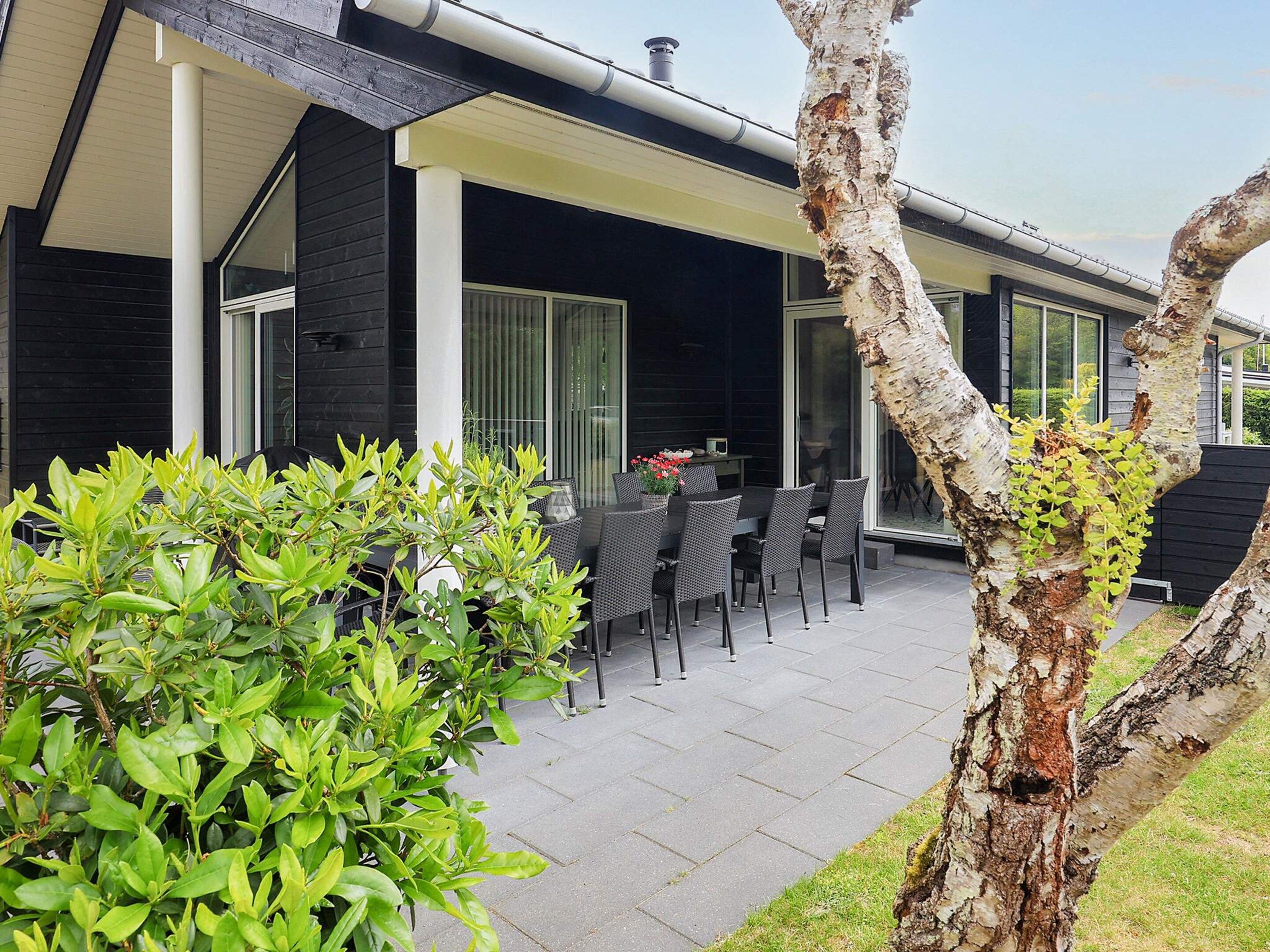 A modern outdoor patio area with a dining table and chairs, surrounded by greenery and a tree.