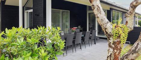 A modern outdoor patio area with a dining table and chairs, surrounded by greenery and a tree.