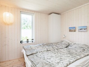 A room with a bed, a window with a view of greenery, a wall-mounted shelf with decorative items, and a wall with two framed pictures.