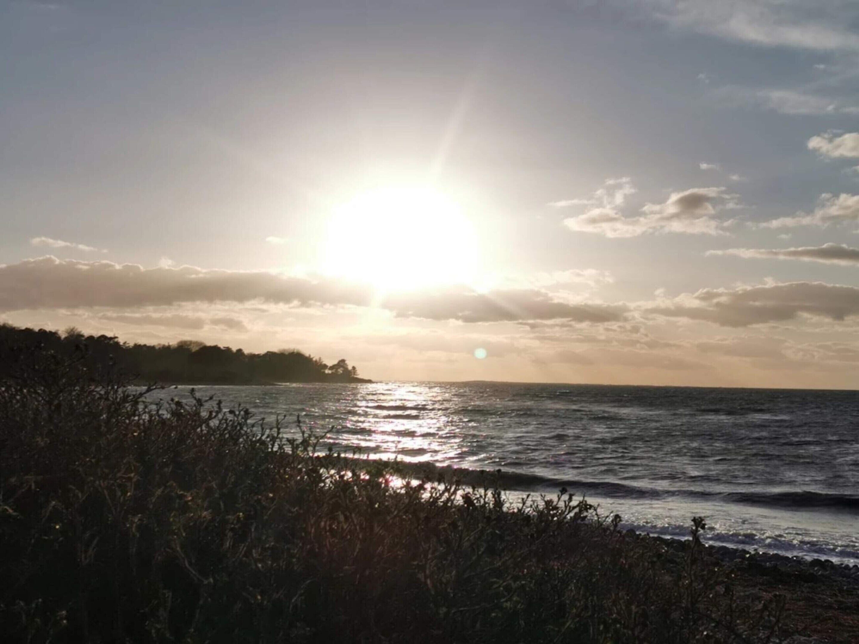 En strandscen med solen som går ner över havet, en avlägsen kustlinje med vegetation och en klar himmel.