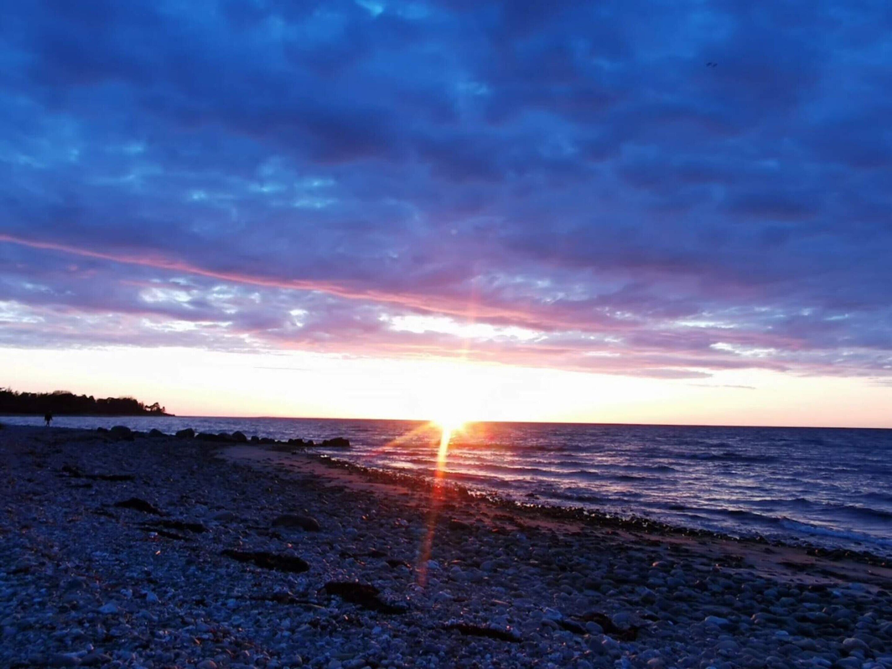 Ranta auringonlaskun aikaan, kivikkoisella rannalla ja kirkas taivas.