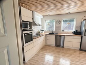 A modern kitchen with white cabinetry, a built-in oven, microwave, and dishwasher. There are two windows with blinds, a wooden ceiling, and a clock on the wall. - 4 Star Holiday Home in Storvorde (Storvorde)
