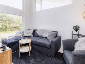Sala de estar moderna com sofá cinza, mesa de madeira redonda e vista para o verde lá fora.