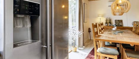 A modern kitchen with a stainless steel refrigerator, a wooden dining table with chairs, and a hanging light fixture.