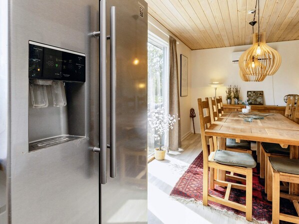 A modern kitchen with a stainless steel refrigerator, a wooden dining table with chairs, and a hanging light fixture.