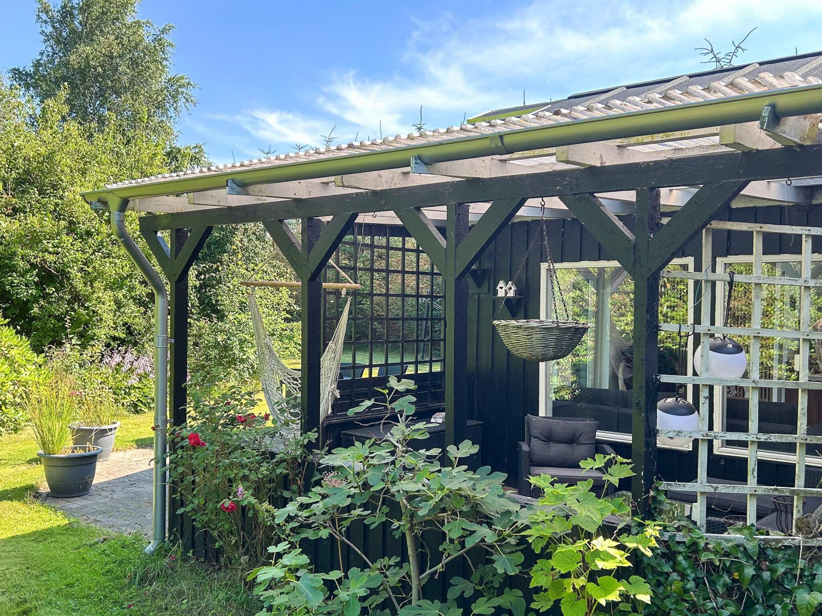 Puinen pergola, jossa on laattakatto, roikkuva amppeli ja punottu tuoli.