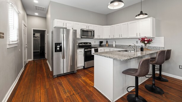 A modern kitchen with white cabinets, stainless steel appliances, a wooden floor, and a bar area with stools. - Historic CozySuites 4BR 2BA  (Louisville)