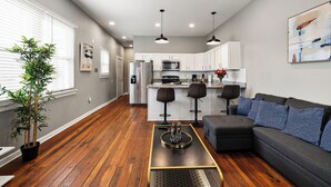 A modern kitchen with white cabinets, a stainless steel refrigerator, and a microwave. There is a dining area with a black table and two bar stools. - Historic CozySuites 4BR 2BA  (Louisville)
