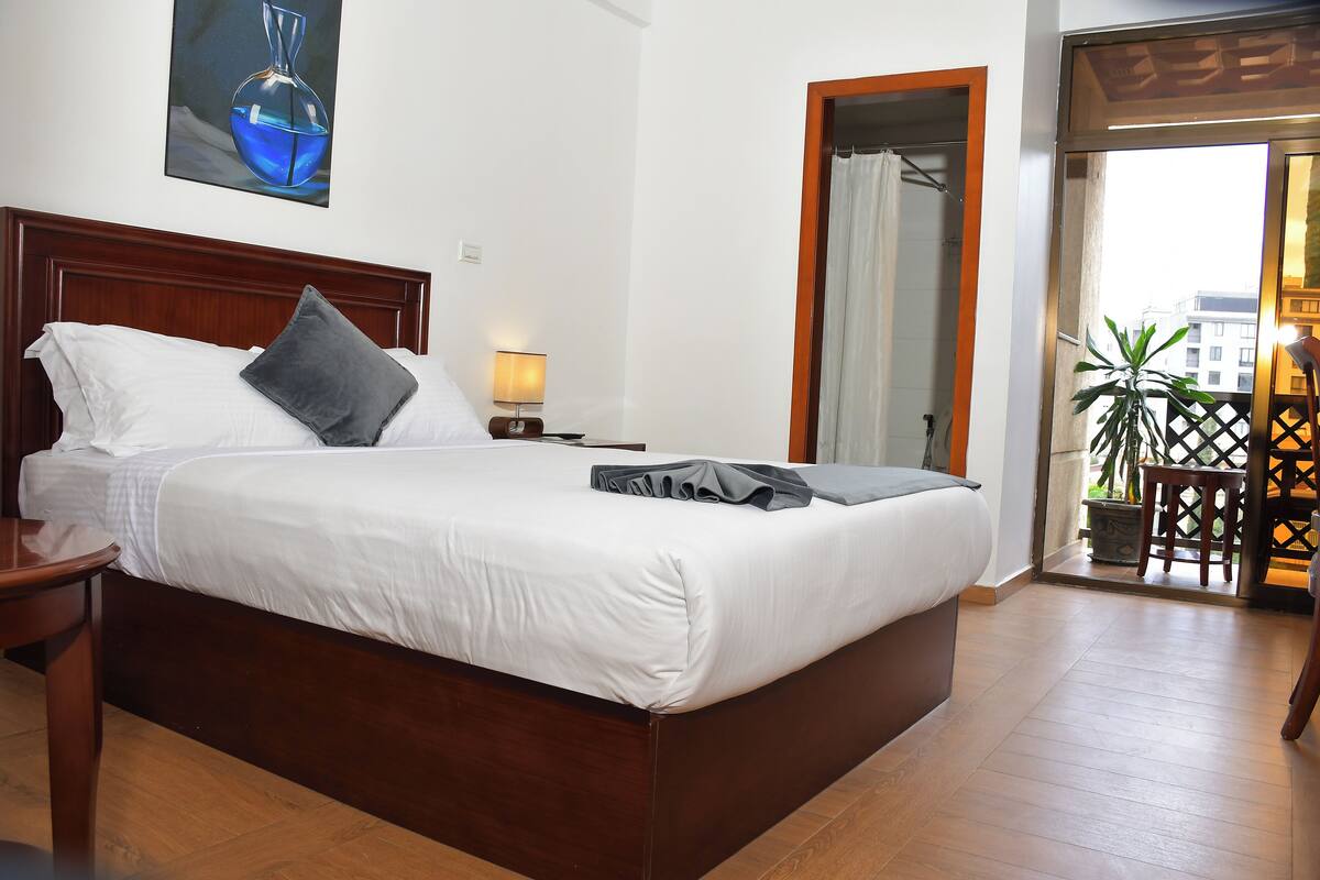 A neatly made bed with white linens and a dark pillow, a wooden nightstand with a lamp, and a view of the outside through a glass door.