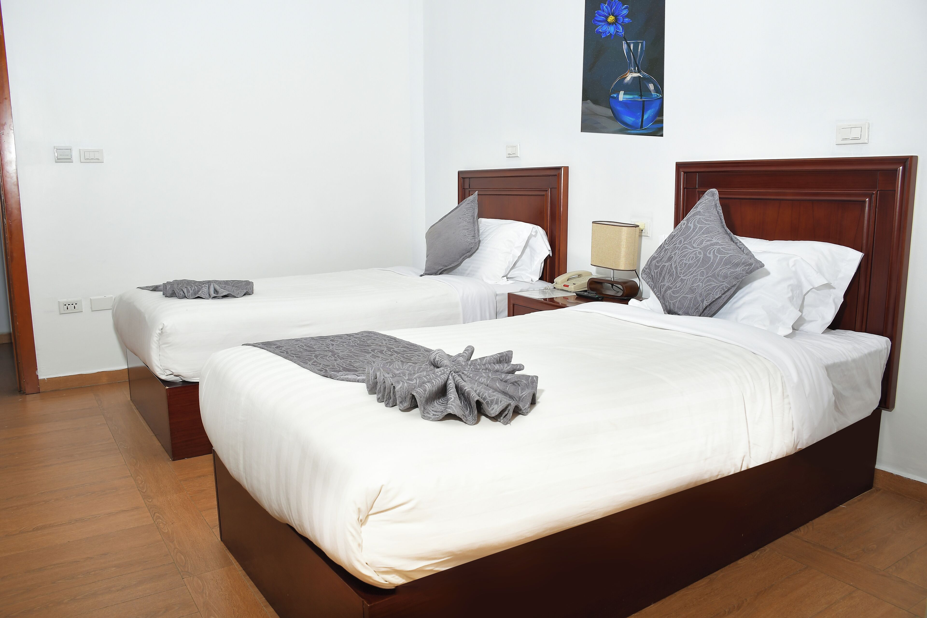 A hotel room with two single beds, each with a wooden headboard, white bedding, and a gray throw blanket. There is a nightstand with a lamp and a telephone.