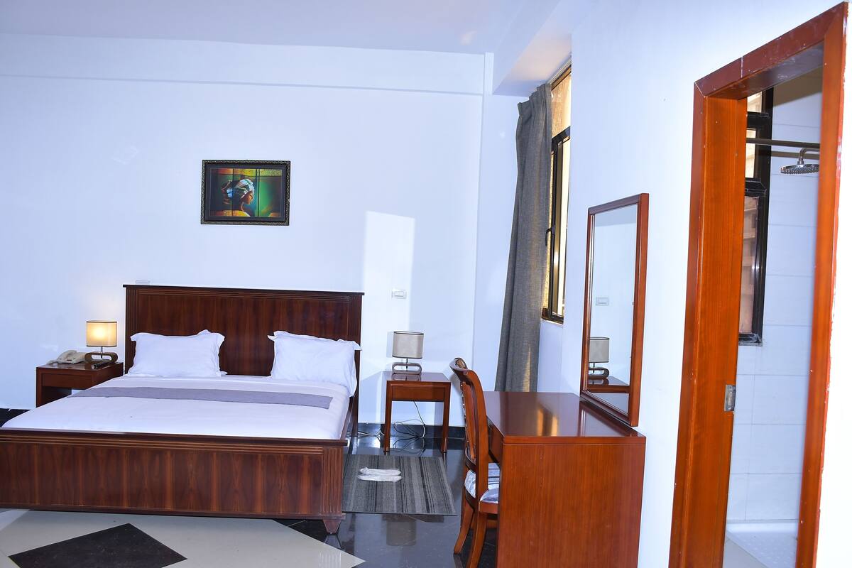 A hotel room with a wooden bed, a desk, a chair, a nightstand with a lamp, and a painting on the wall.