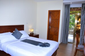 Habitación de hotel con una cama grande, cabecera de madera, mesita de noche con lámpara y vista a un espacio exterior a través de una puerta de vidrio.