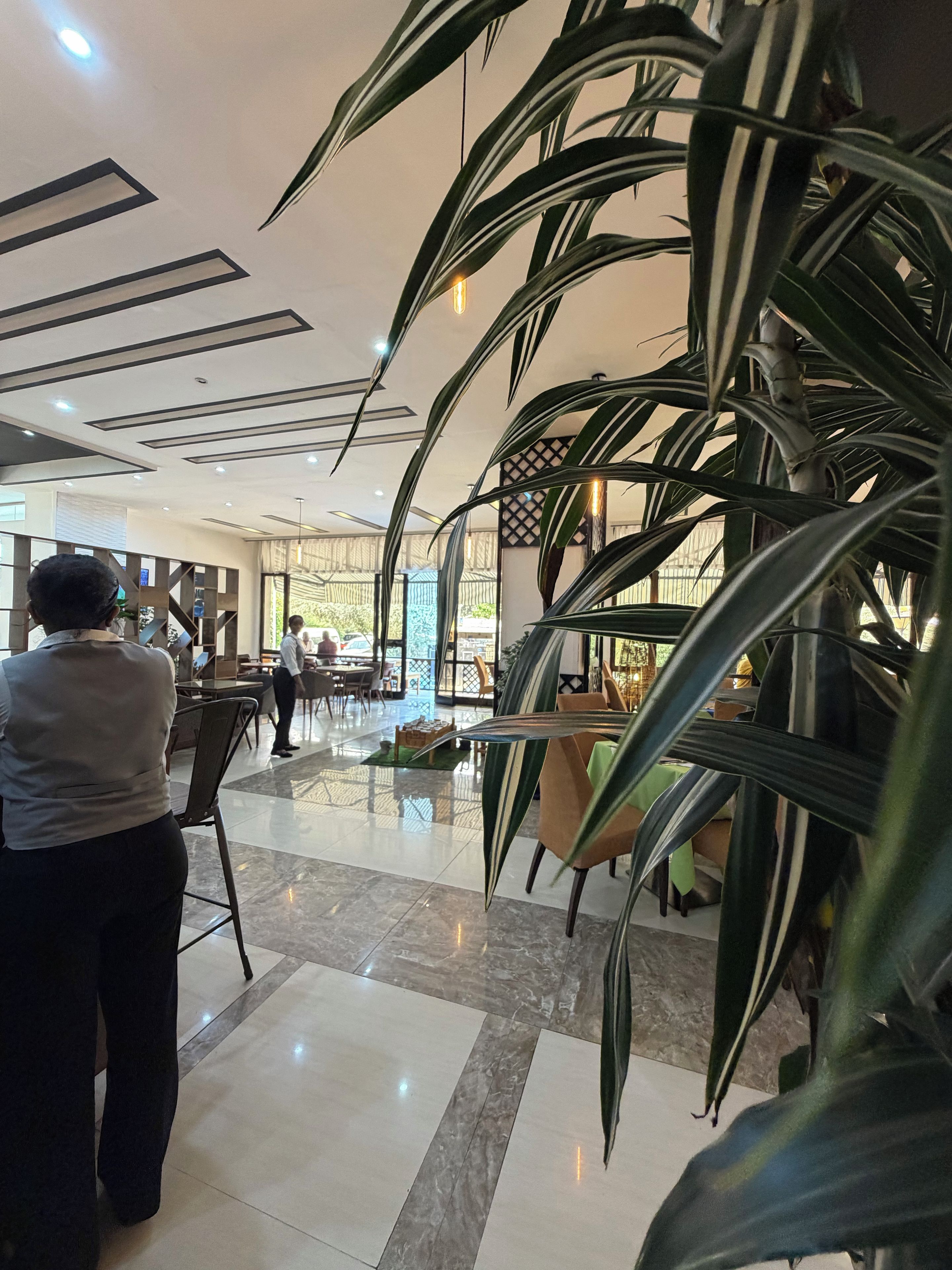 A hotel lobby with a modern design, featuring a marble floor, large potted plants, and a seating area with tables and chairs.