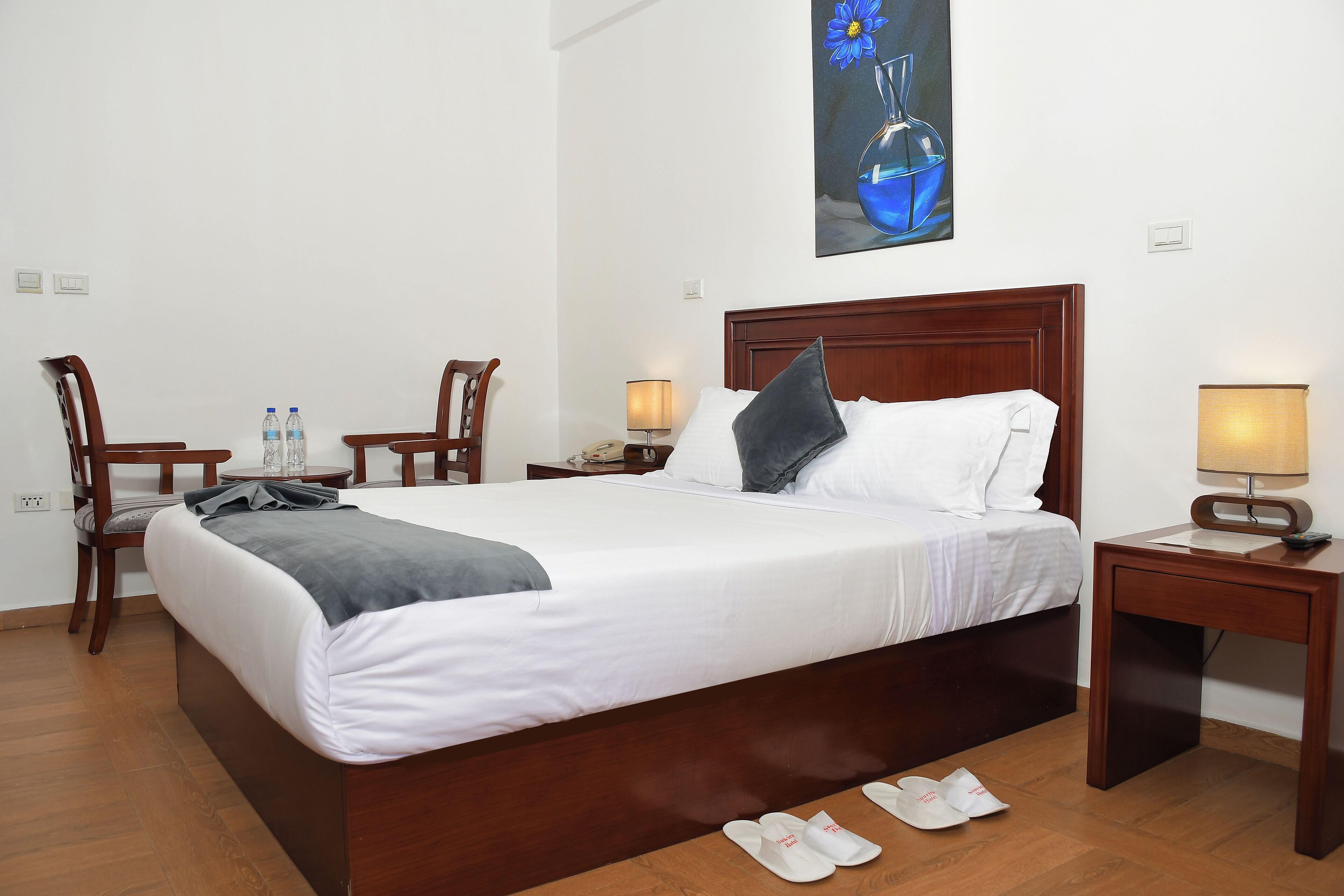 Un lit bien fait, avec du linge de lit blanc et un plaid gris, deux chaises en bois, une petite table avec des bouteilles d’eau et un tableau représentant un vase bleu accroché au mur.