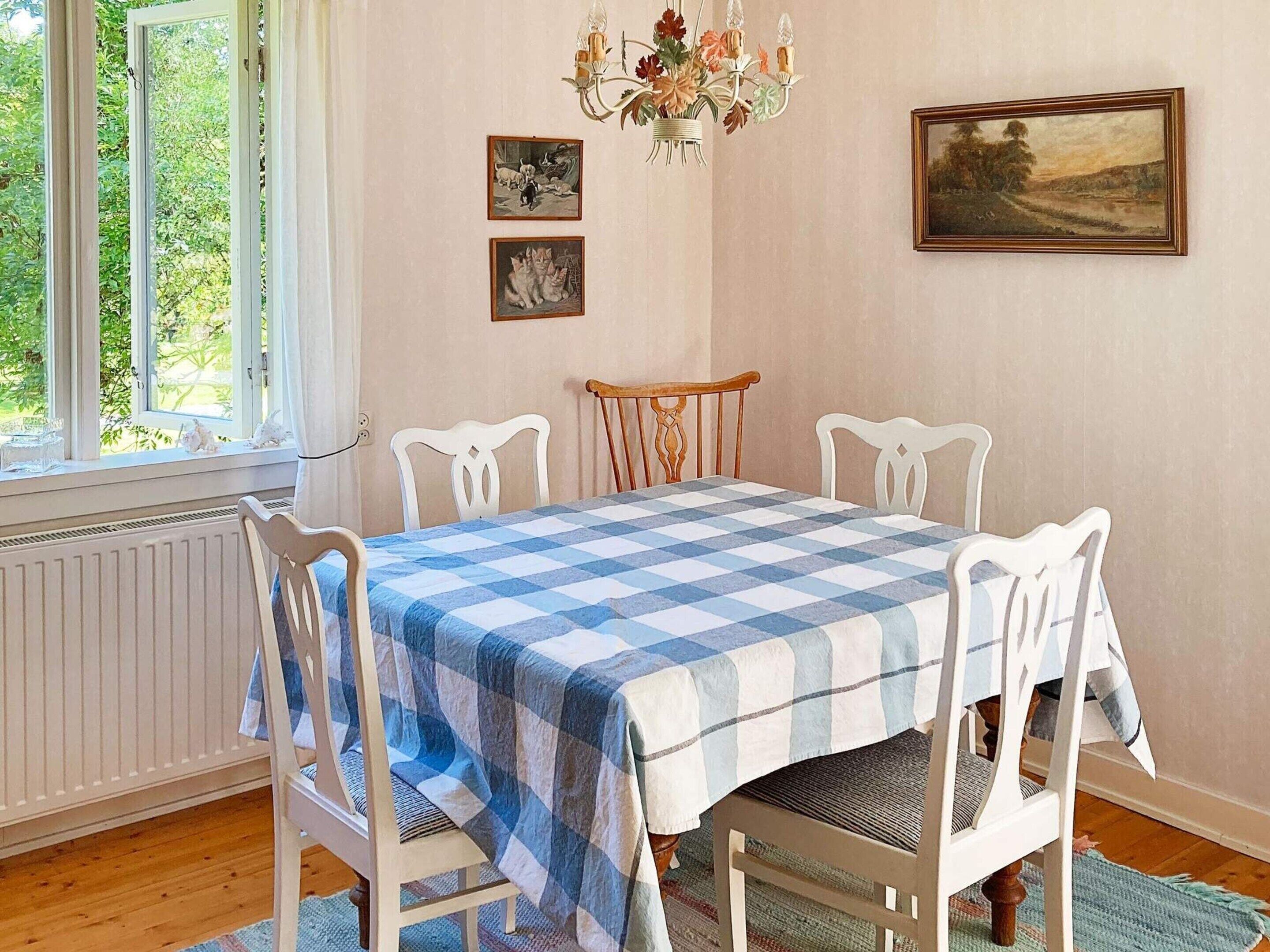 Ett matrum med ett rutigt bordsduksmönster, vita stolar, en kristallkrona, inramade tavlor och ett fönster med utsikt över grönskan.