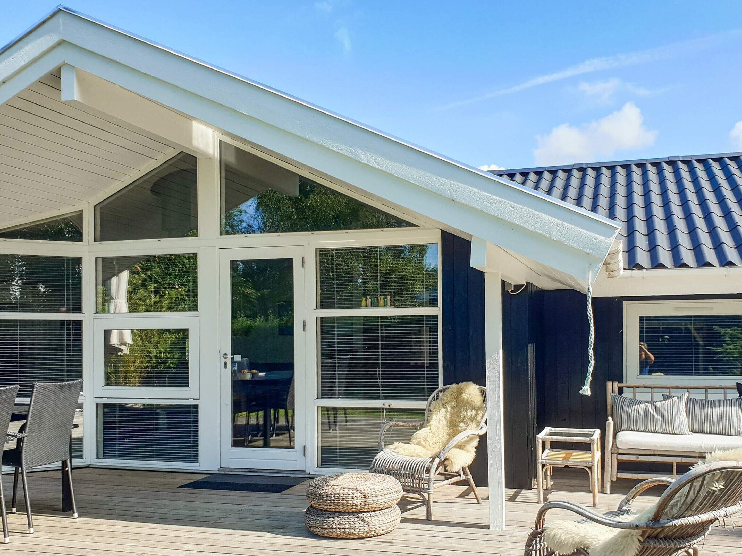 A covered outdoor patio with wicker furniture, a wooden deck, and a house with large glass doors and a tiled roof.