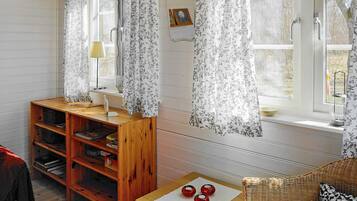 A room with a wooden bookshelf, a small table with red spherical objects, a wicker chair, and a window with floral curtains.